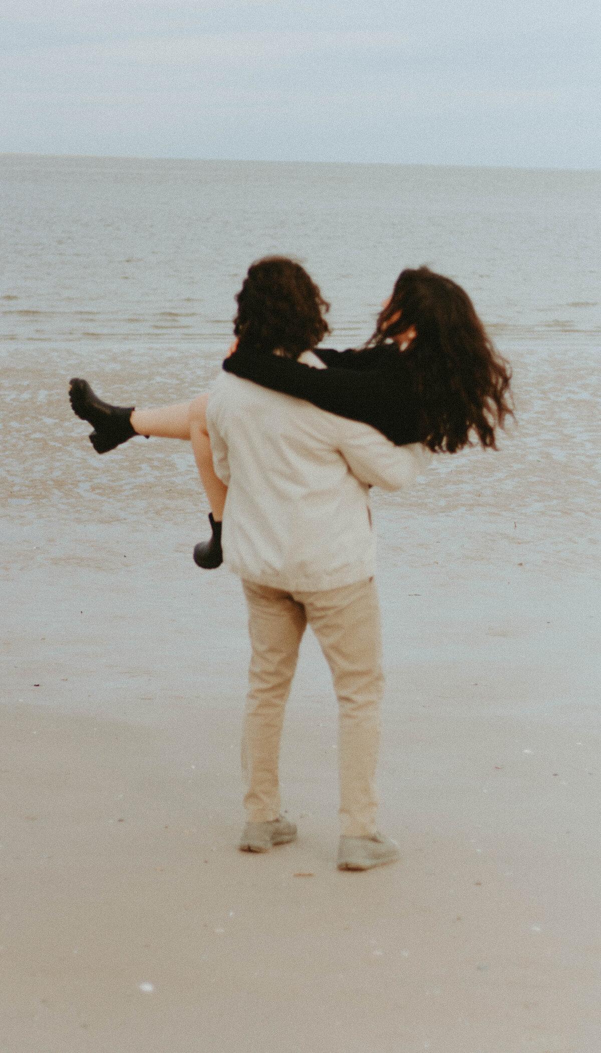 Partner carrying another partner on a wide empty beach at low tide, soft film tones