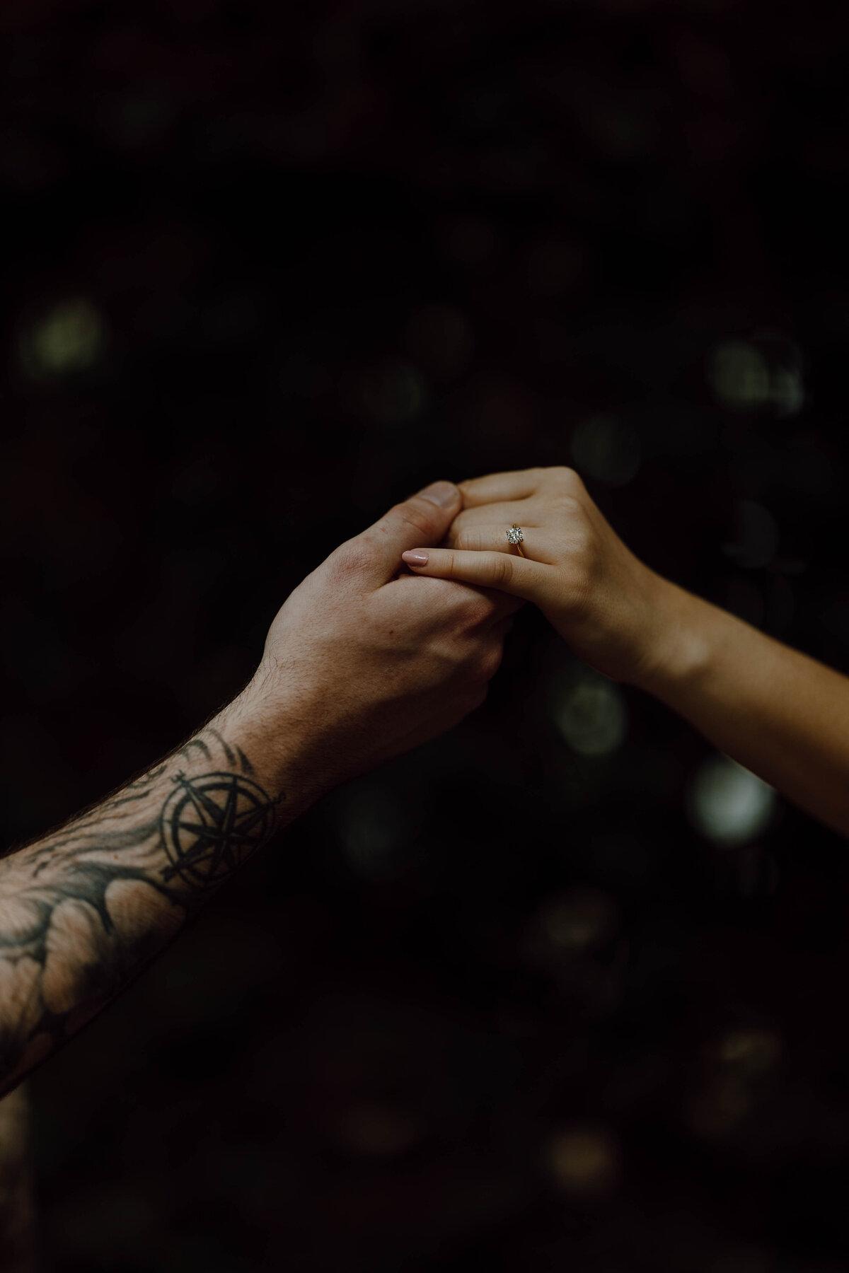 Two hands clasped in low light, a solitaire engagement ring catching the edge of the frame