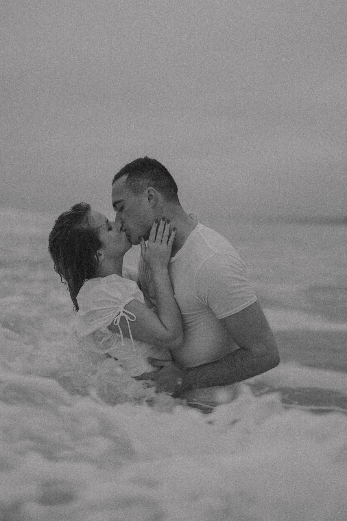 Couple kissing waist-deep in ocean waves, moody black and white, editorial