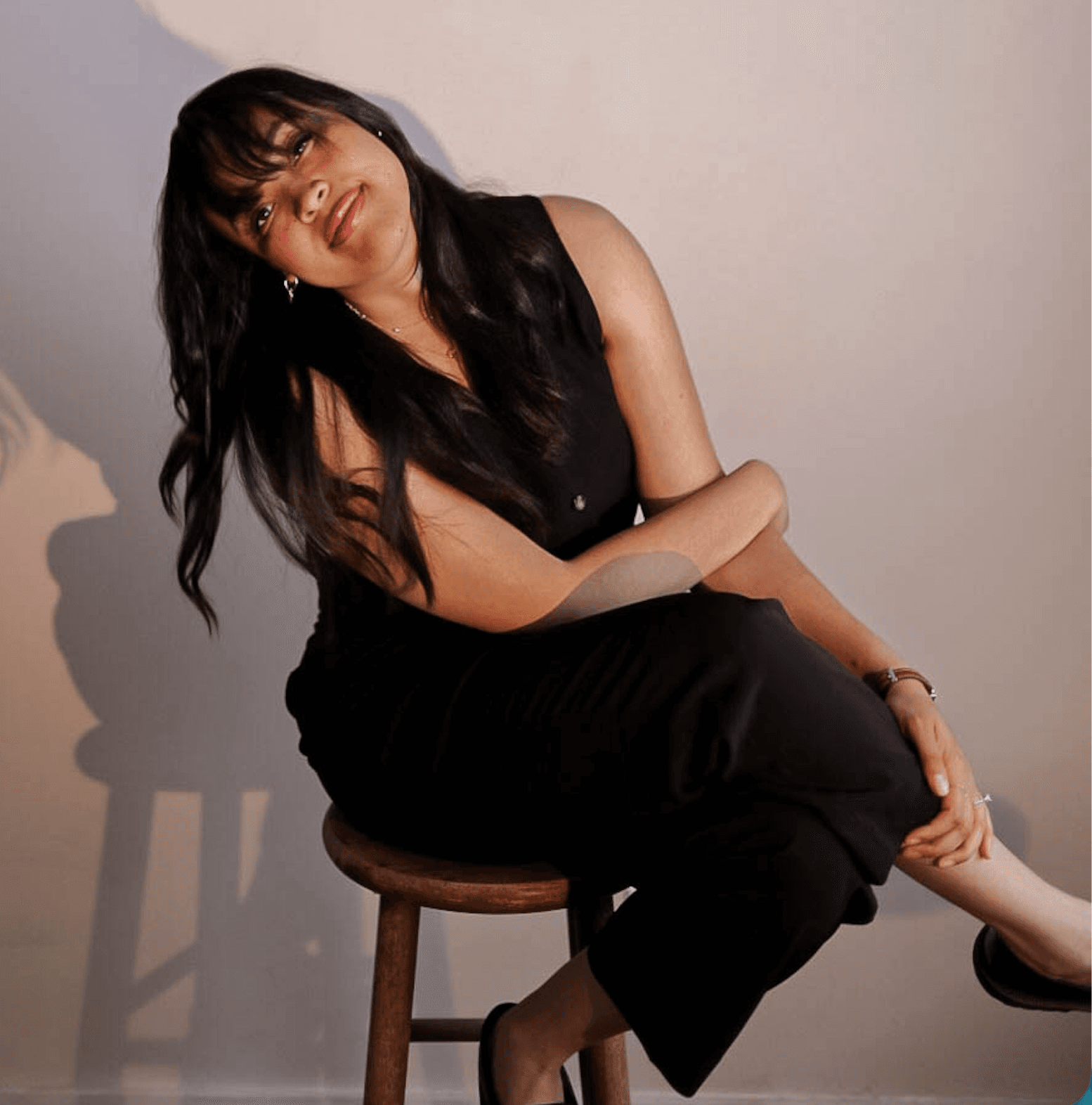 Hallel Saine seated on a studio stool in a black dress, soft cast shadow on a warm cream backdrop