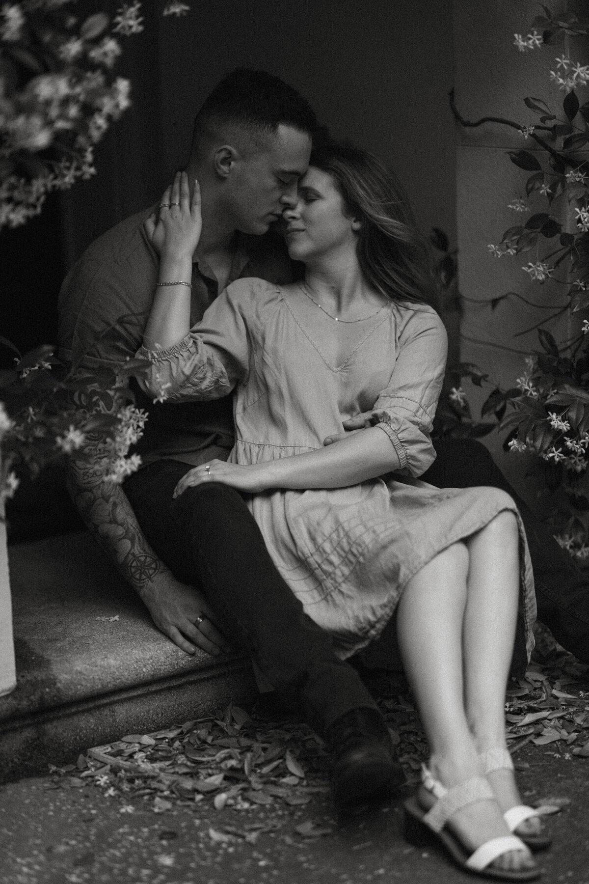 Couple seated on stoop, moody black-and-white portrait framed by jasmine flowers